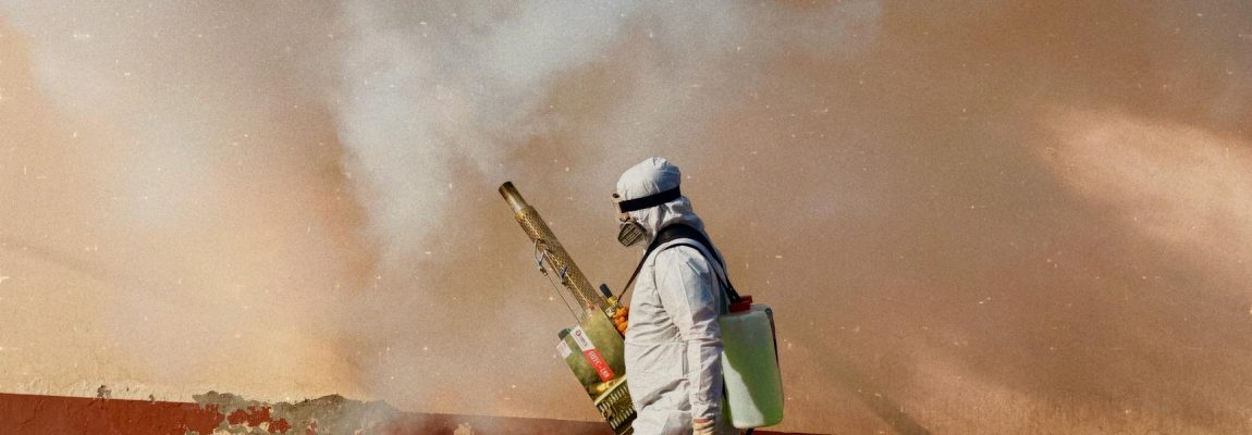 A pest control worker in a yellow uniform and respirator sprays pesticide inside a room.